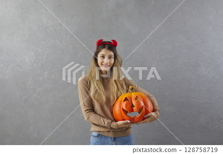 Portrait of happy young woman or teenage girl in devil horns holding Halloween pumpkin 128758719