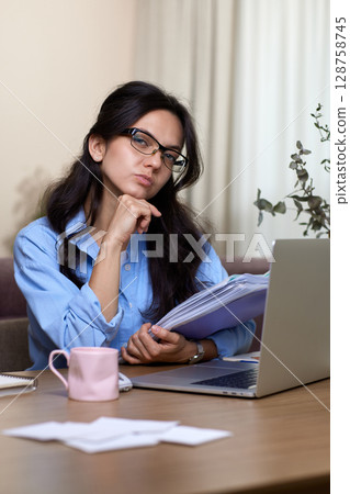 portrait of woman in blue shirt holds folders uses laptop in home office 128758745