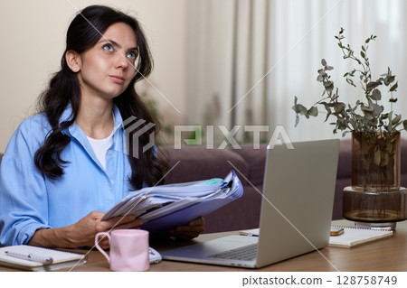 portrait of woman in blue shirt holds folders uses laptop in home office 128758749