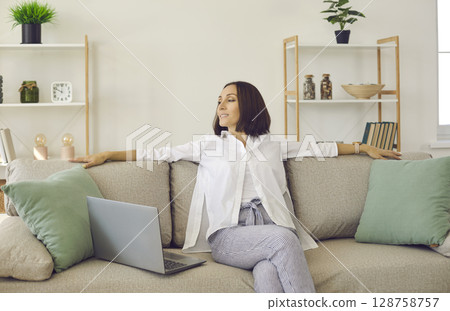 Smiling and calm adult woman sitting on sofa near laptop computer relaxing at home. 128758757