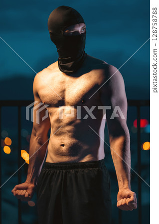 Muscular athlete with mask posing powerfully at night, showcasing strength and mystery, Toned man with face covering displaying power and intensity in evening silhouette 128758788