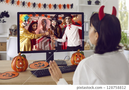 Woman waving hello at computer screen during virtual Halloween costume party with friends 128758812