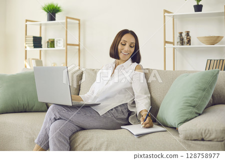 Woman writes in notebook while sitting on sofa at home and listening to online webinar on laptop. 128758977