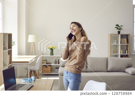 Joyful young woman receives good news during pleasant conversation on mobile phone at home. Joyful young woman receives good news during pleasant conversation on mobile phone at home. 128758984