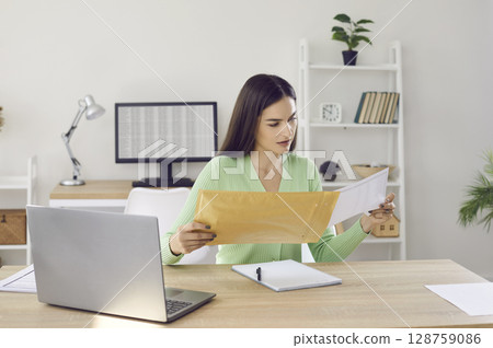 Young woman holding envelope and reading letter while sitting at desk with laptop at home 128759086