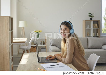 Happy teen girl in headphones at home preparing homework sitting with notebook at laptop. Happy teen girl in headphones at home preparing homework sitting with notebook at laptop. 128759216