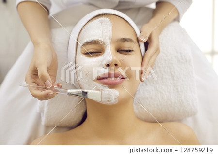 Happy young woman enjoying cosmetic face mask during facial procedure in spa salon 128759219