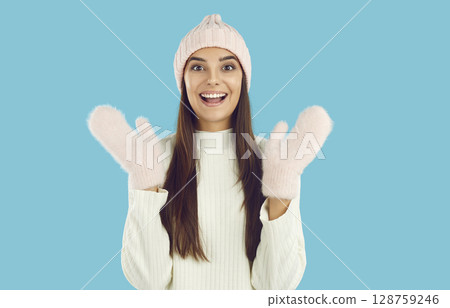 Portrait of happy girl in pink hat, mittens and white sweater waving hands on blue background. Portrait of happy girl in pink hat, mittens and white sweater waving hands on blue background. 128759246