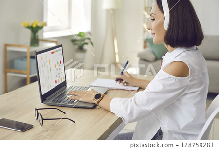 Woman sitting in front of laptop at home, studying online business course and taking notes 128759281