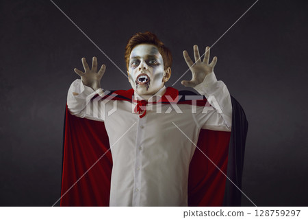 Studio portrait of boy wearing spooky Halloween makeup, fake fangs and vampire cape Studio portrait of boy wearing spooky Halloween makeup, fake fangs and vampire cape 128759297