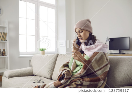 Woman who has flu or cold sitting on sofa, looking at thermometer and calling doctor Woman who has flu or cold sitting on sofa, looking at thermometer and calling doctor 128759317
