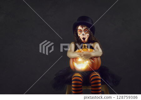 Shocked little girl in Halloween clothes sitting with jack-o-lantern on black background. Shocked little girl in Halloween clothes sitting with jack-o-lantern on black background. 128759386