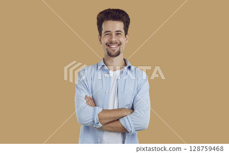 Portrait of smiling young man with neatly shaved beard and mustache, dressed in white T-shirt and blue shirt, confidently looking straight, arms crossed on his chest. 128759468