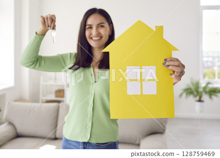 Happy single young woman smiling and holding paper house and keys to her new home 128759469