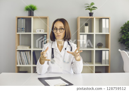 Video call portrait of professional physician sitting at office desk and talking Video call portrait of professional physician sitting at office desk and talking 128759512