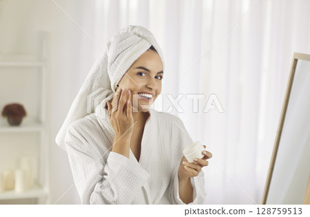 Portrait of beautiful young woman taking care of her skin on her face while standing in bathroom. Portrait of beautiful young woman taking care of her skin on her face while standing in bathroom. 128759513