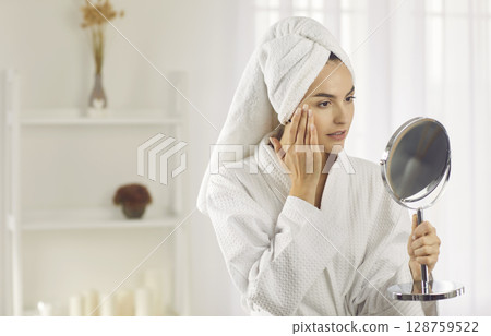 Beautiful young woman in white towel looking in mirror during her skin care routine Beautiful young woman in white towel looking in mirror during her skin care routine 128759522