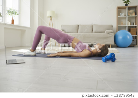 Young woman doing glute bridge exercise at home while watching online workout video 128759542