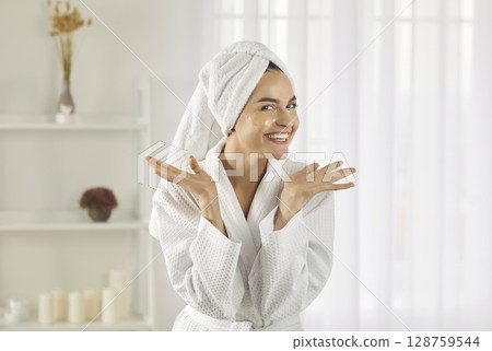 Happy woman wearing bathrobe and towel on her head performing her daily skin care routine at home. 128759544