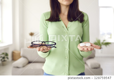 Young woman is holding glasses and eye lenses and trying to choose which to wear Young woman is holding glasses and eye lenses and trying to choose which to wear 128759547