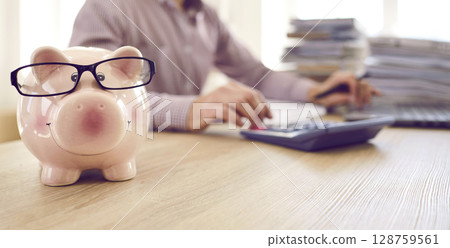 Banner with piggy bank wearing glasses on table of accountant who's working in background 128759561