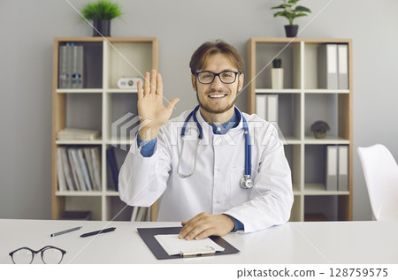 Webcam portrait of happy online doctor sitting at desk and waving hand at camera Webcam portrait of happy online doctor sitting at desk and waving hand at camera 128759575