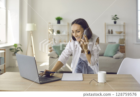 Friendly senior female entrepreneur having business conversation on mobile phone in her home office. Friendly senior female entrepreneur having business conversation on mobile phone in her home office. 128759576