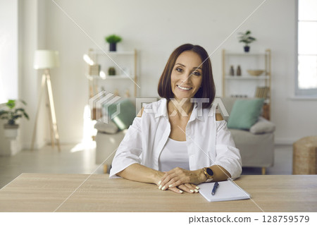 Happy smiling middle aged woman in casual shirt sitting at desk with notebook at home Happy smiling middle aged woman in casual shirt sitting at desk with notebook at home 128759579