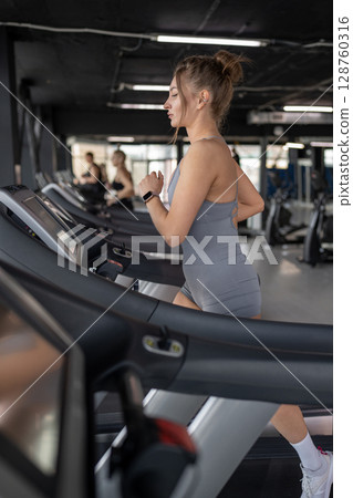 A young woman runs on a treadmill in a gym, showing her dedication to fitness and wellbeing A young woman runs on a treadmill in a gym, showing her dedication to fitness and wellbeing 128760316