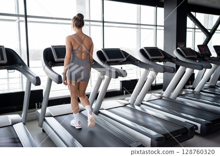 A fit woman confidently walking on a treadmill in a bright and inviting gym, promoting fitness and overall health. 128760320