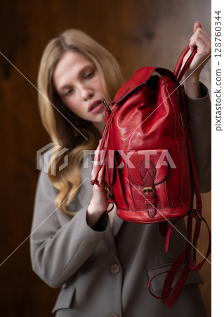 A stylish young woman, long blonde hair, poses in a grey suit, holding a vibrant red leather backpack against a warm wooden background A stylish young woman, long blonde hair, poses in a grey suit, holding a vibrant red leather backpack against a warm wooden background 128760344