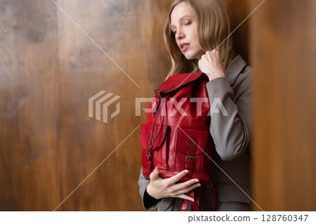 A stylish young woman, long blonde hair, poses in a grey suit, holding a vibrant red leather backpack against a warm wooden background A stylish young woman, long blonde hair, poses in a grey suit, holding a vibrant red leather backpack against a warm wooden background 128760347