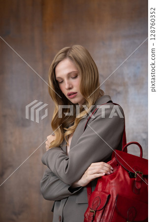 A stylish young woman, long blonde hair, poses in a grey suit, holding a vibrant red leather backpack against a warm wooden background A stylish young woman, long blonde hair, poses in a grey suit, holding a vibrant red leather backpack against a warm wooden background 128760352