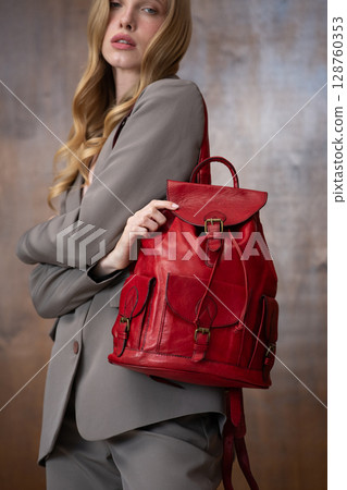 A stylish young woman, long blonde hair, poses in a grey suit, holding a vibrant red leather backpack against a warm wooden background A stylish young woman, long blonde hair, poses in a grey suit, holding a vibrant red leather backpack against a warm wooden background 128760353