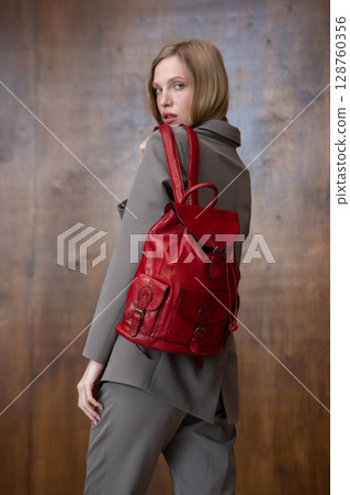 A stylish young woman, long blonde hair, poses in a grey suit, holding a vibrant red leather backpack against a warm wooden background A stylish young woman, long blonde hair, poses in a grey suit, holding a vibrant red leather backpack against a warm wooden background 128760356