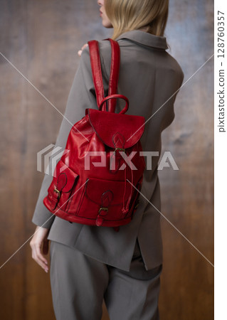 A stylish young woman, long blonde hair, poses in a grey suit, holding a vibrant red leather backpack against a warm wooden background A stylish young woman, long blonde hair, poses in a grey suit, holding a vibrant red leather backpack against a warm wooden background 128760357