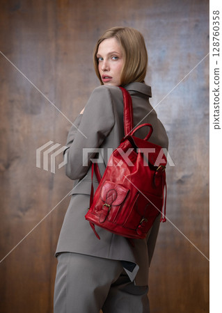 A stylish young woman, long blonde hair, poses in a grey suit, holding a vibrant red leather backpack against a warm wooden background A stylish young woman, long blonde hair, poses in a grey suit, holding a vibrant red leather backpack against a warm wooden background 128760358