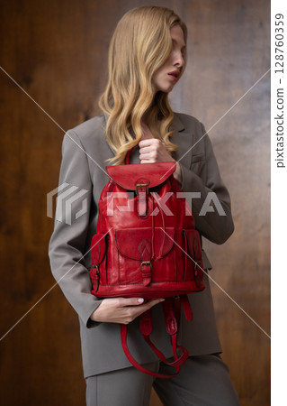 A stylish young woman, long blonde hair, poses in a grey suit, holding a vibrant red leather backpack against a warm wooden background A stylish young woman, long blonde hair, poses in a grey suit, holding a vibrant red leather backpack against a warm wooden background 128760359