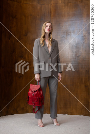 A stylish young woman, poses in a grey suit, holding a vibrant yellow leather backpack against a warm wooden background 128760360
