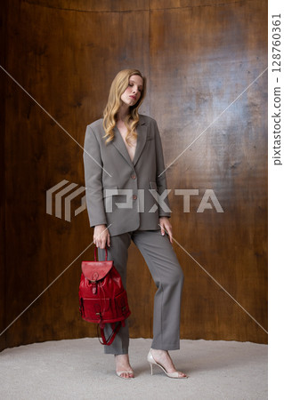 A stylish young woman, long blonde hair, poses in a grey suit, holding a vibrant red leather backpack against a warm wooden background 128760361