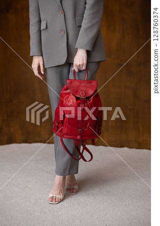 A stylish young woman, poses in a grey suit, holding a vibrant red leather backpack against a warm wooden background A stylish young woman, poses in a grey suit, holding a vibrant red leather backpack against a warm wooden background 128760374