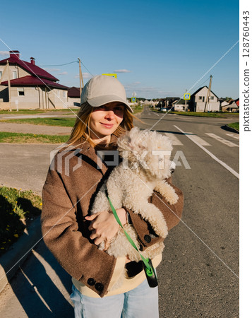 Young woman wearing casual clothes and baseball cap holding small white dog in arms, standing in suburban street with residential houses and asphalt road, during sunny day. Woman holding toy poodle Young woman wearing casual clothes and baseball cap holding small white dog in arms, standing in suburban street with residential houses and asphalt road, during sunny day. Woman holding toy poodle 128760443