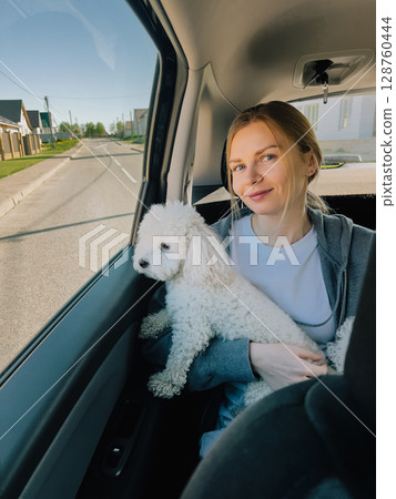 Young woman traveling in back seat of car, cradling her small poodle dog in her arms while smiling and looking directly at camera, enjoying sunny road trip adventure. Young woman cuddling small poodle Young woman traveling in back seat of car, cradling her small poodle dog in her arms while smiling and looking directly at camera, enjoying sunny road trip adventure. Young woman cuddling small poodle 128760444