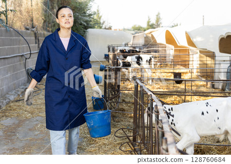 Asian woman holds bucket filled with water and waters calves in street cattle pen. 128760684