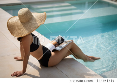 Woman relaxing by poolside, wearing large sunhat, reading magazine, enjoying summer leisure time 128761715