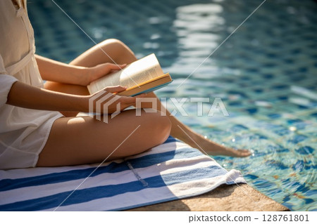 Woman relaxing by the poolside reading a book on a sunny day with water reflections in background 128761801