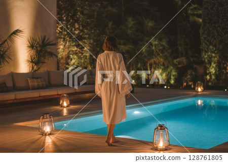 Woman in white robe walking by illuminated poolside at night with tranquil ambiance and lush greenery 128761805