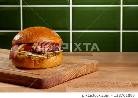 Delicious burger with bacon, cheese, and pickles resting on wooden cutting board 128761896
