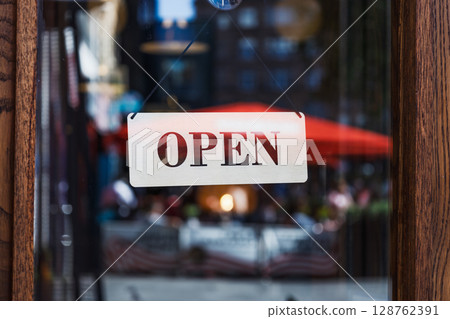 Open Sign on Storefront Glass with Blurred City Street and Outdoor Cafe Reflections 128762391