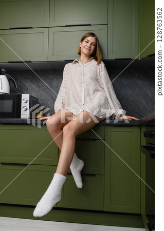 Relaxed young woman sitting on kitchen counter in cozy outfit. Relaxed young woman sitting on kitchen counter in cozy outfit. 128763562